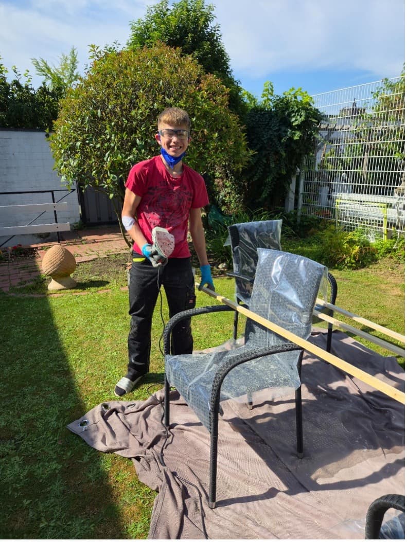 Jugendlicher mit Schutzbrille und Handschuhen beim Schleifen eines Gartenstuhls im sonnigen Garten
